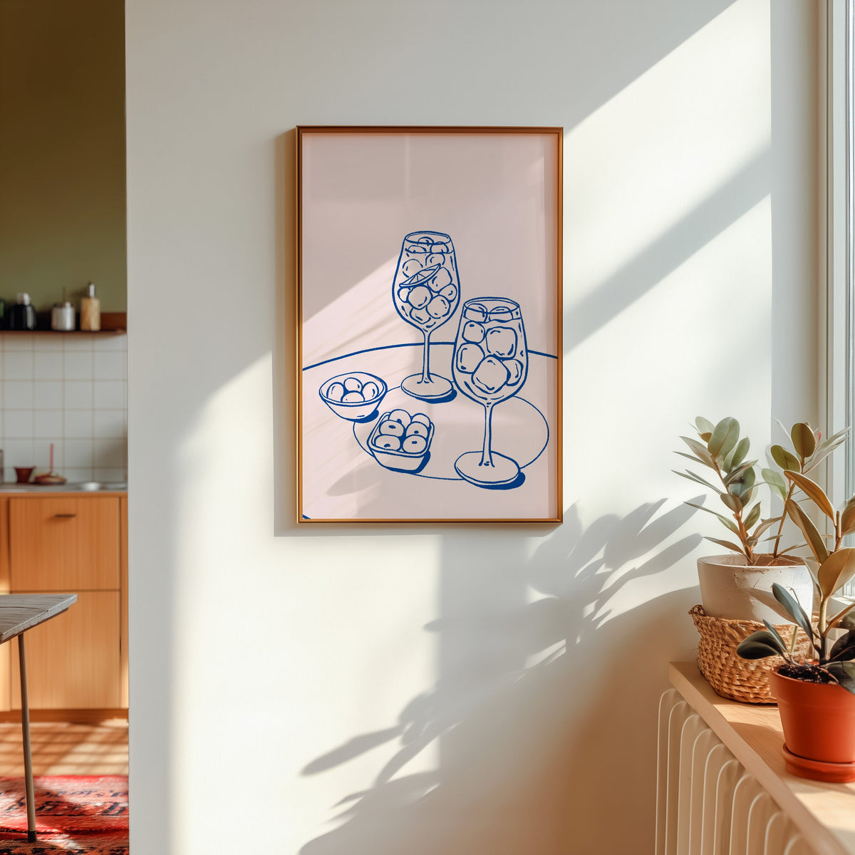 Spritz and olives print in a gold frame in sunlight with plants and a kitchen beyond