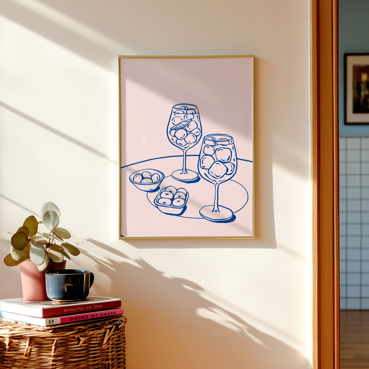 Spritz and olives print in a gold frame above a basket of books and a pilea plant
