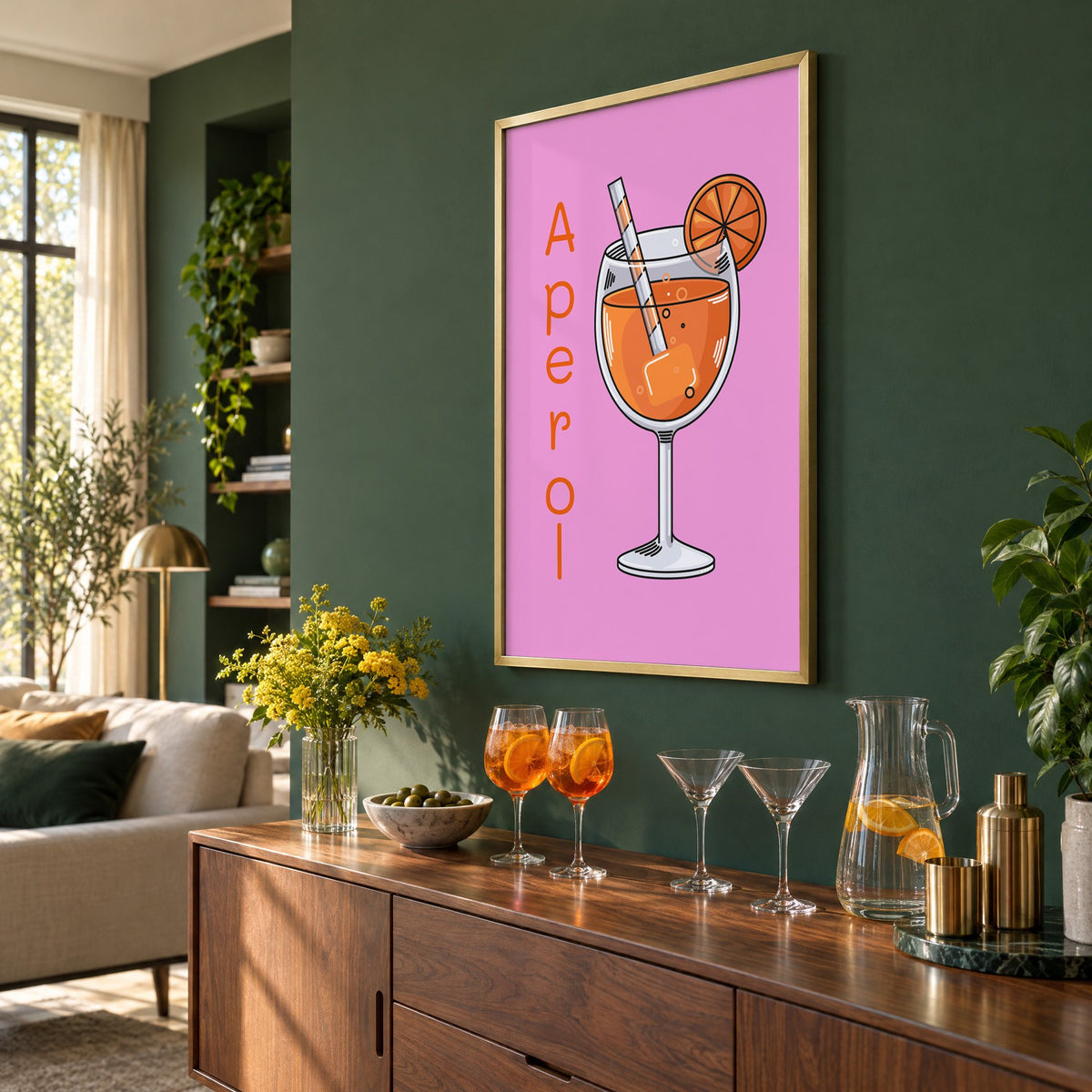 Orange spritz glass on pink print in a brass frame on a bottle green wall above a sideboard with spritz and martini glasses, seen at an angle