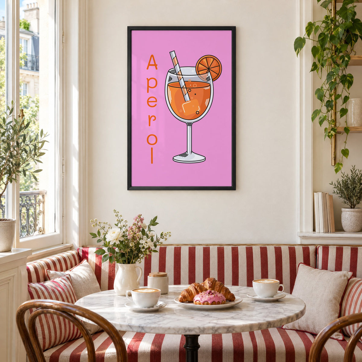 Orange spritz glass on pink print in a black frame above a striped cafe breakfast nook with cappuccino and pastries
