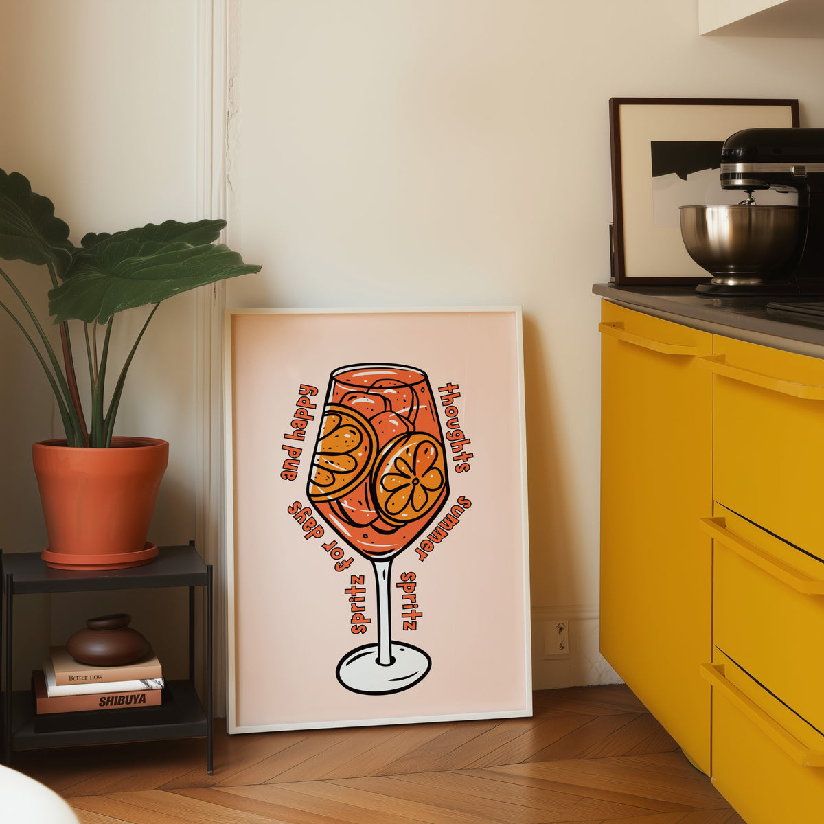 Spritz glass print in a white frame leaning on parquet beside a bright yellow kitchen
