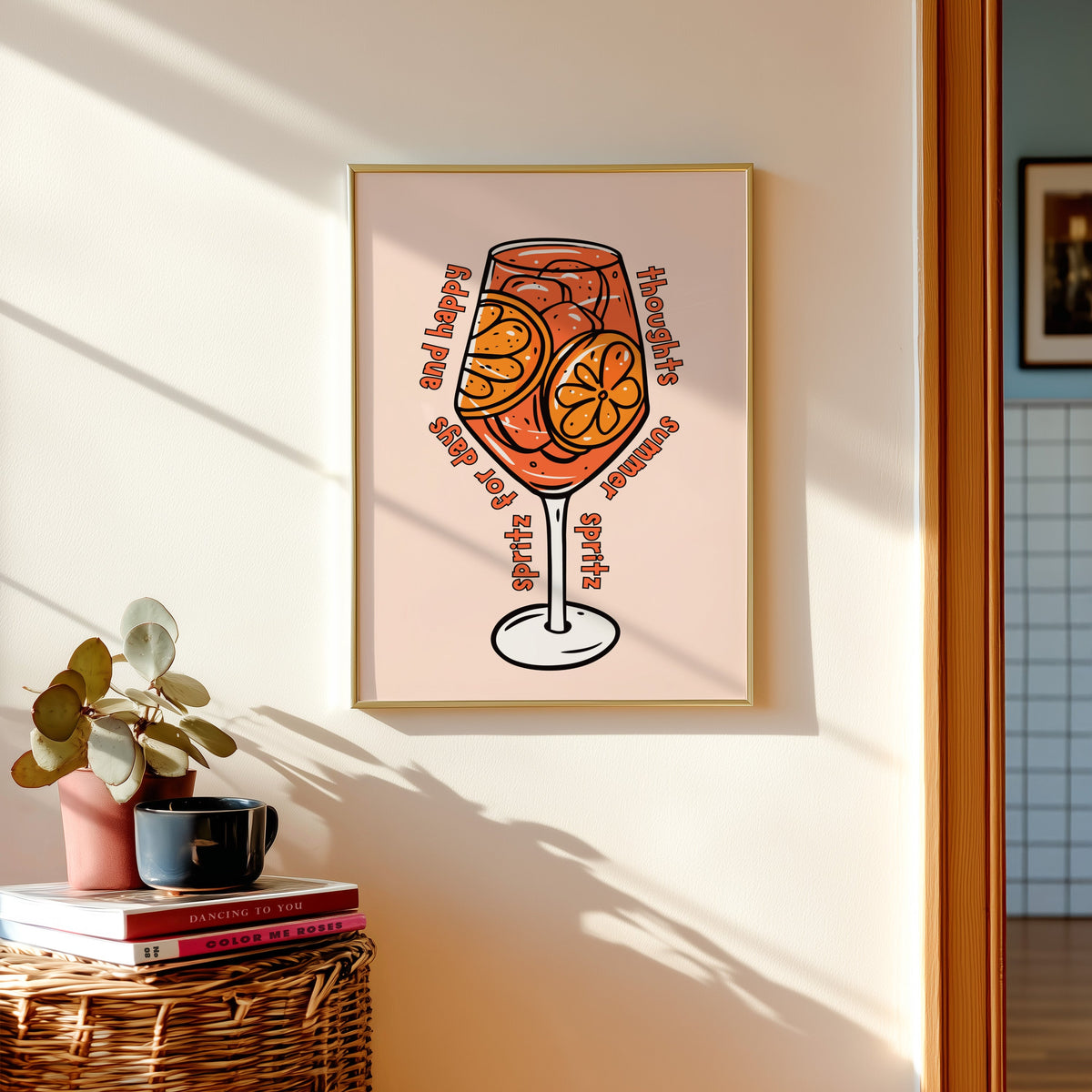Spritz glass print in a gold frame above a basket of books and a pilea plant