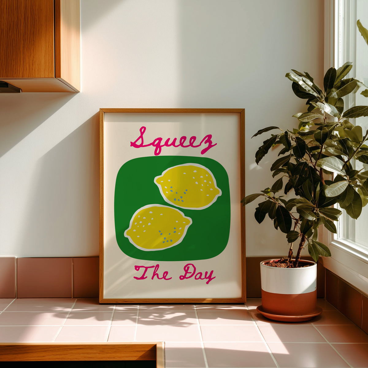 Squeez the Day lemons print in a golden oak frame leaning on a pink tiled ledge in strong afternoon sun