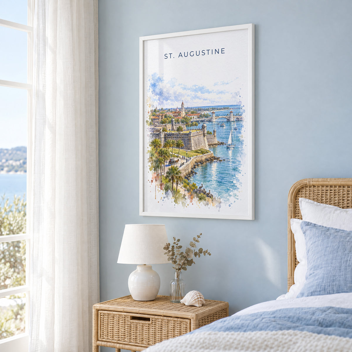 St. Augustine watercolor print in a white frame on a pale blue wall beside a rattan bed in a coastal bedroom