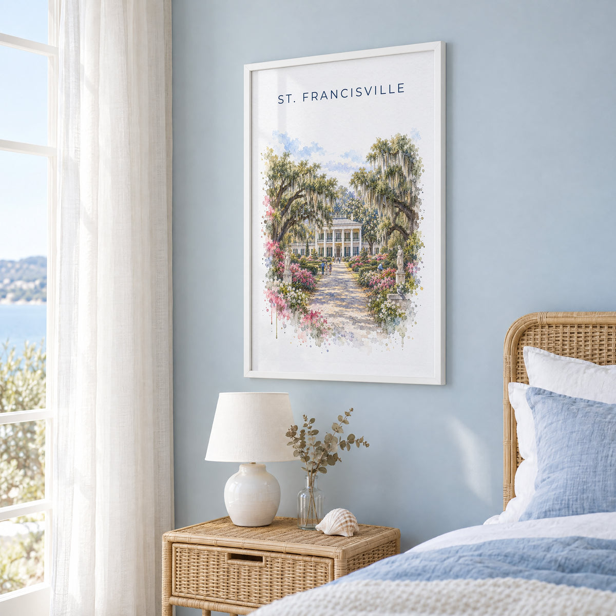St. Francisville, Louisiana watercolor print in a white frame on a pale blue wall beside a rattan bed in a coastal bedroom