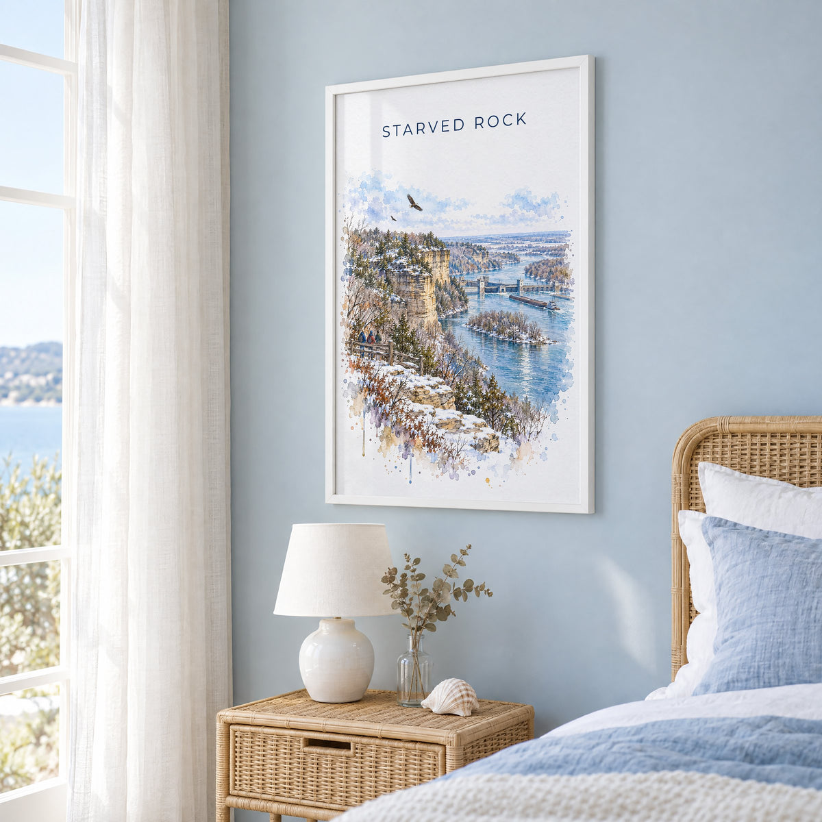 Starved Rock watercolor print in a white frame on a pale blue wall beside a rattan bed in a coastal bedroom