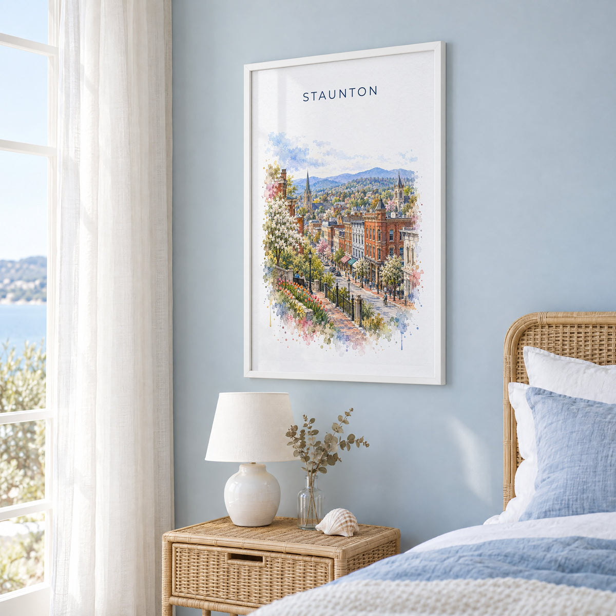 Staunton, Virginia watercolor print in a white frame on a pale blue wall beside a rattan bed in a coastal bedroom