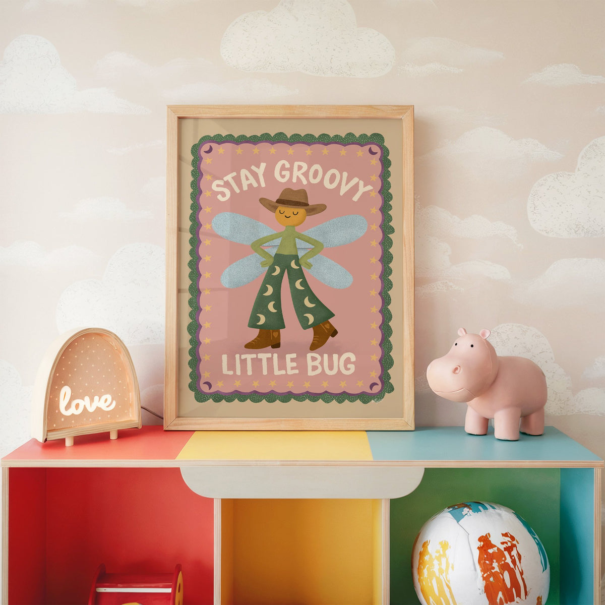 Stay groovy little bug cowboy nursery print in an oak frame on a rainbow toy shelf against cloud wallpaper