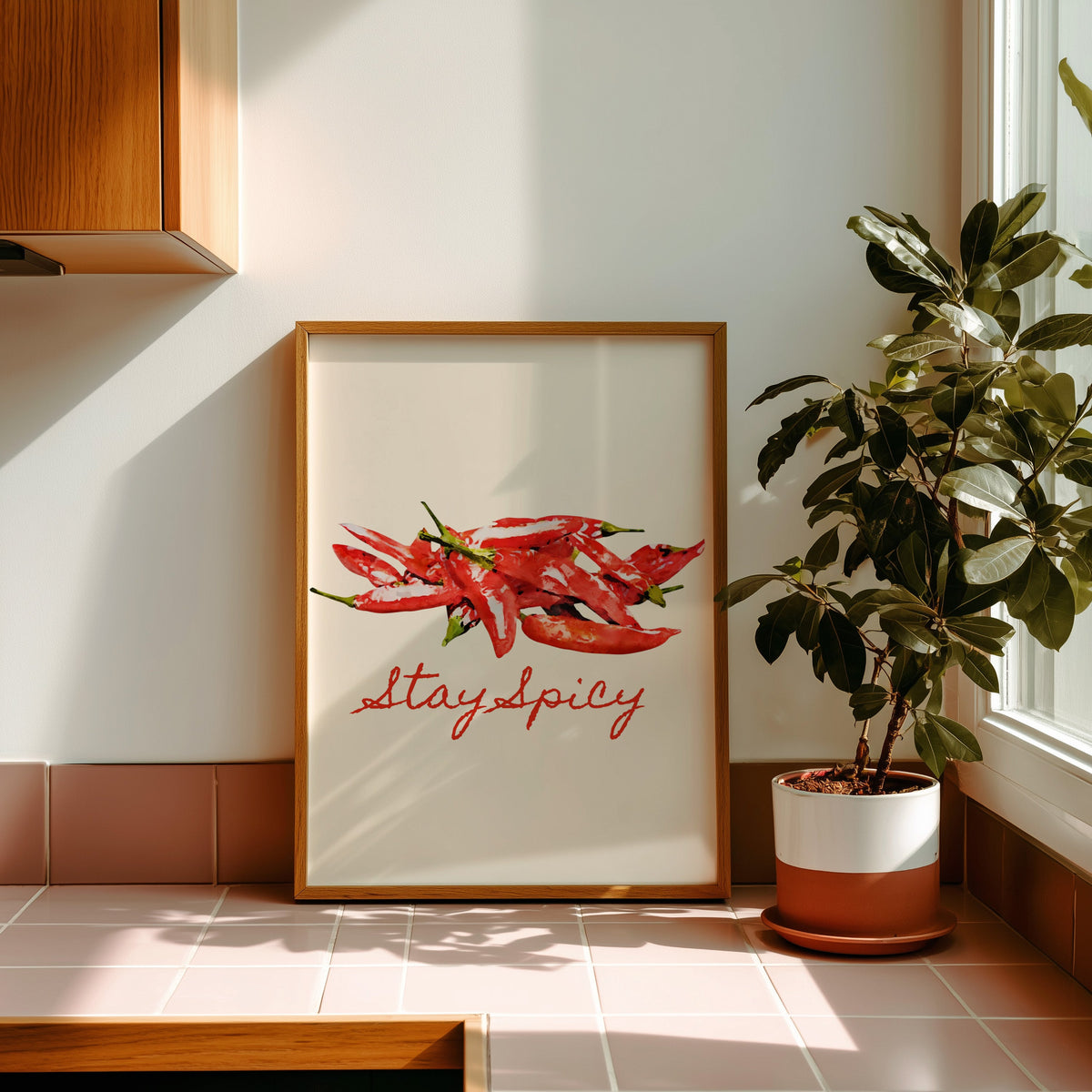 Stay Spicy watercolour chillies print in a golden oak frame leaning on a pink tiled ledge in strong afternoon sun
