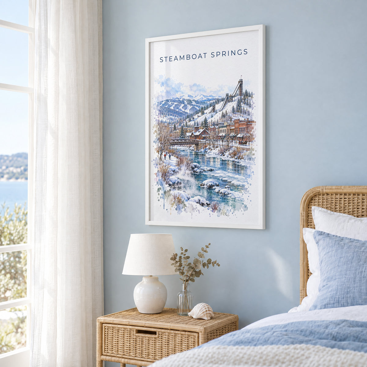 Steamboat Springs watercolor print in a white frame on a pale blue wall beside a rattan bed in a coastal bedroom