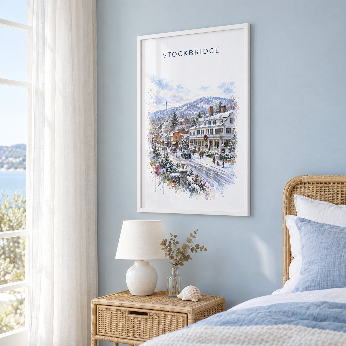Stockbridge, Massachusetts watercolor print in a white frame on a pale blue wall beside a rattan bed in a coastal bedroom