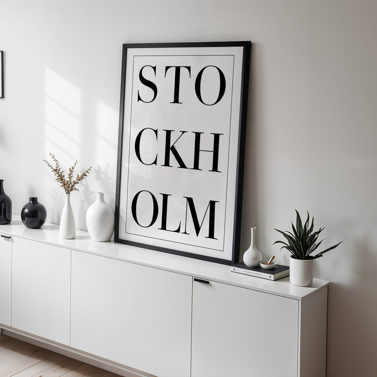 Stockholm stacked serif lettering on white print in a black frame on a white sideboard with black and white vases