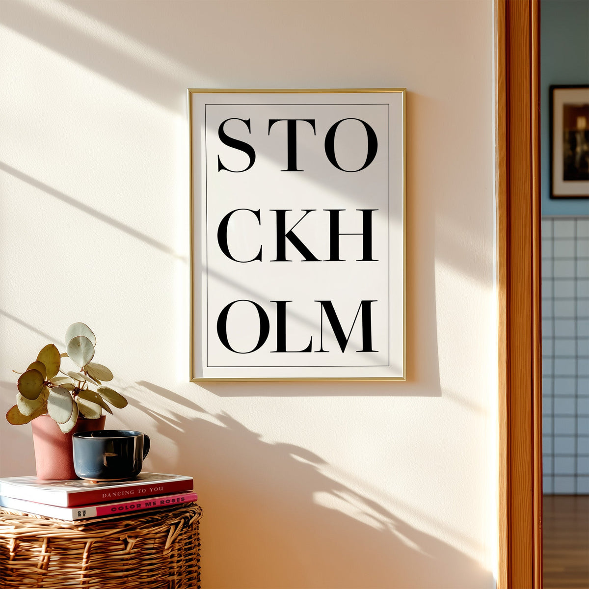 Stockholm stacked serif lettering on white print shown in a gold frame above a wicker basket and stacked books