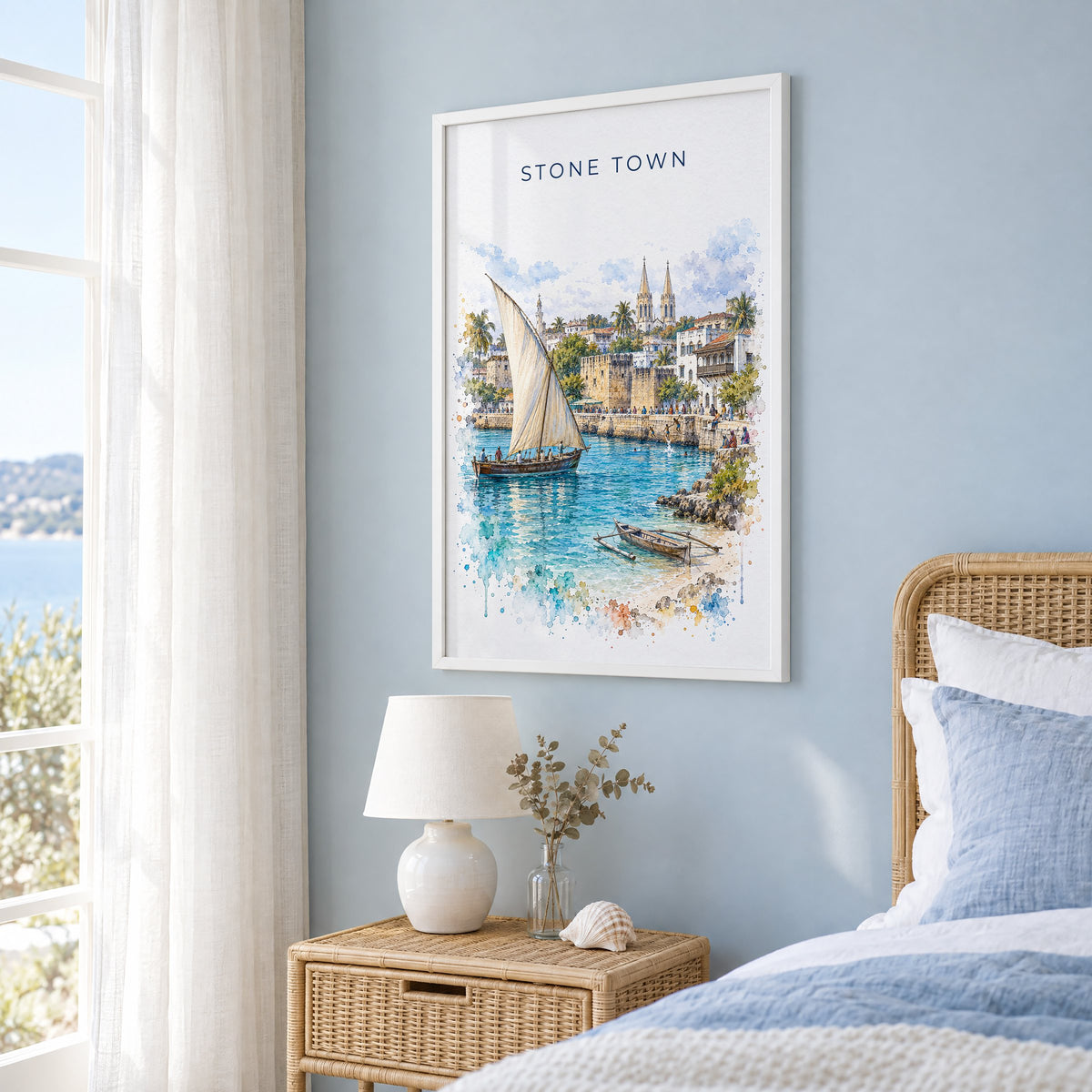Stone Town watercolor print in a white frame on a pale blue wall beside a rattan bed in a coastal bedroom