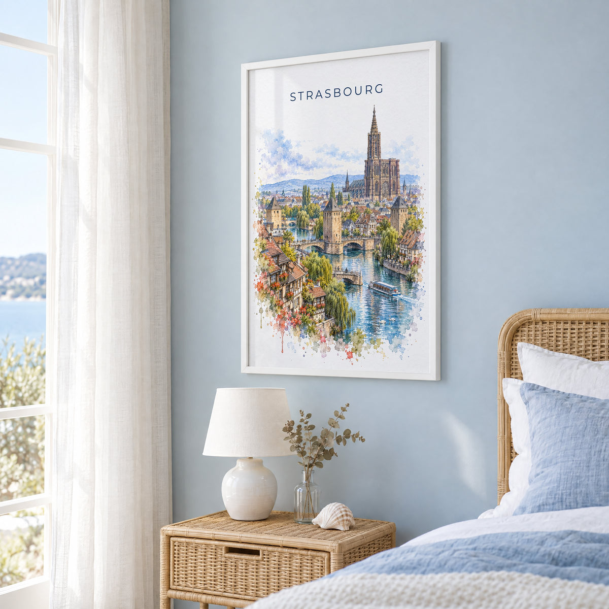 Strasbourg watercolor print in a white frame on a pale blue wall beside a rattan bed in a coastal bedroom