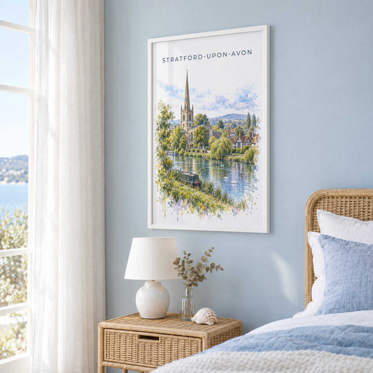 Stratford-upon-Avon watercolor print in a white frame on a pale blue wall beside a rattan bed in a coastal bedroom