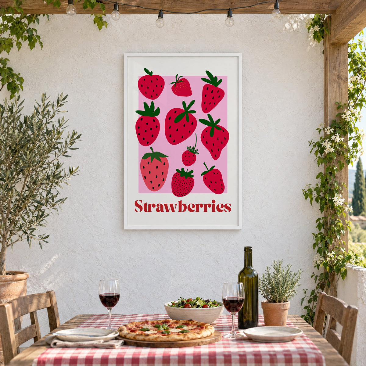 Strawberries bold serif pink panel berry print in a white frame on a stucco wall above a checked-cloth table with pizza and red wine