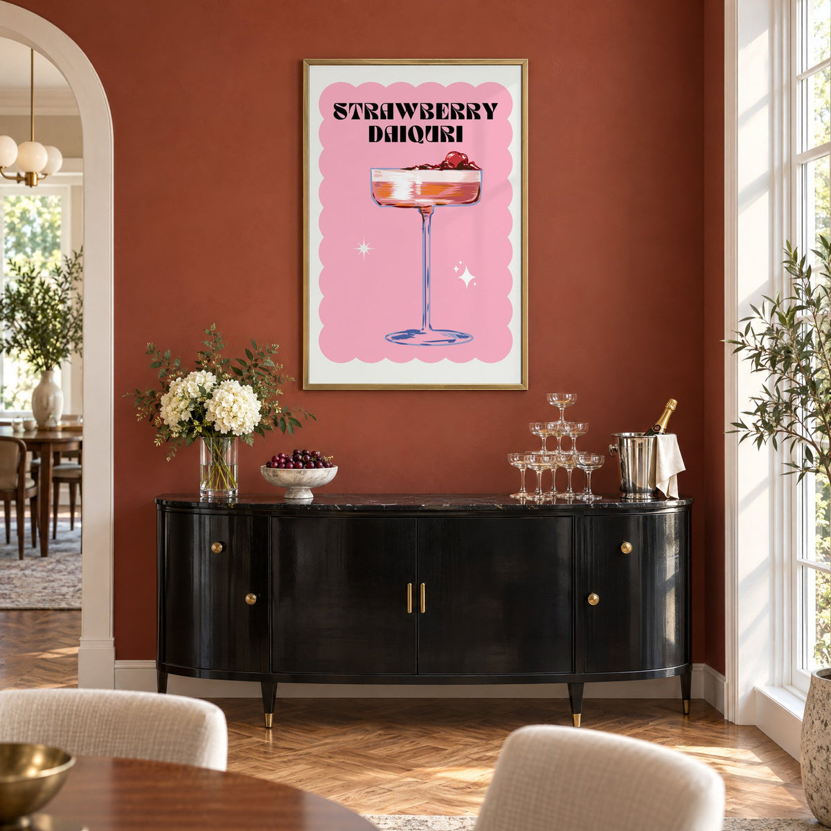 Strawberry daiquiri pink coupe print in a brass frame on a terracotta wall above a black buffet with a champagne tower