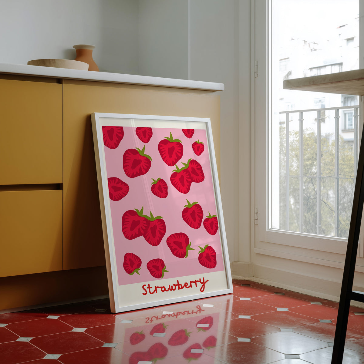 Strawberry print in a white frame against yellow cabinets on a red tiled floor