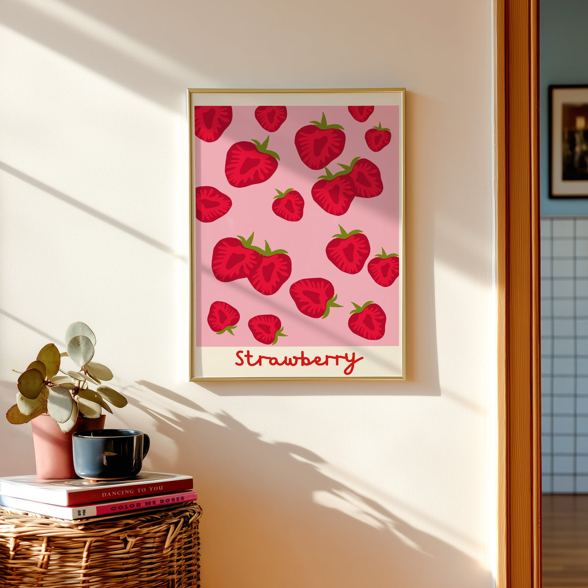 Strawberry print in a gold frame above a basket of books and a pilea plant