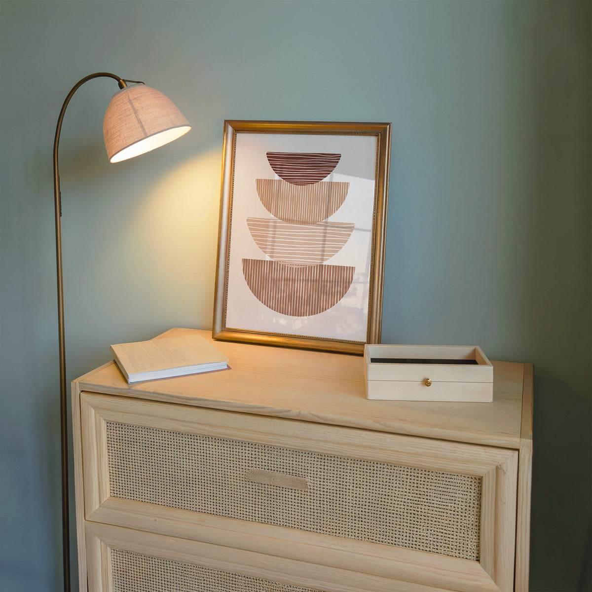 Striped stacked bowls earth tone print shown in a gold frame on a cane dresser lit by a floor lamp
