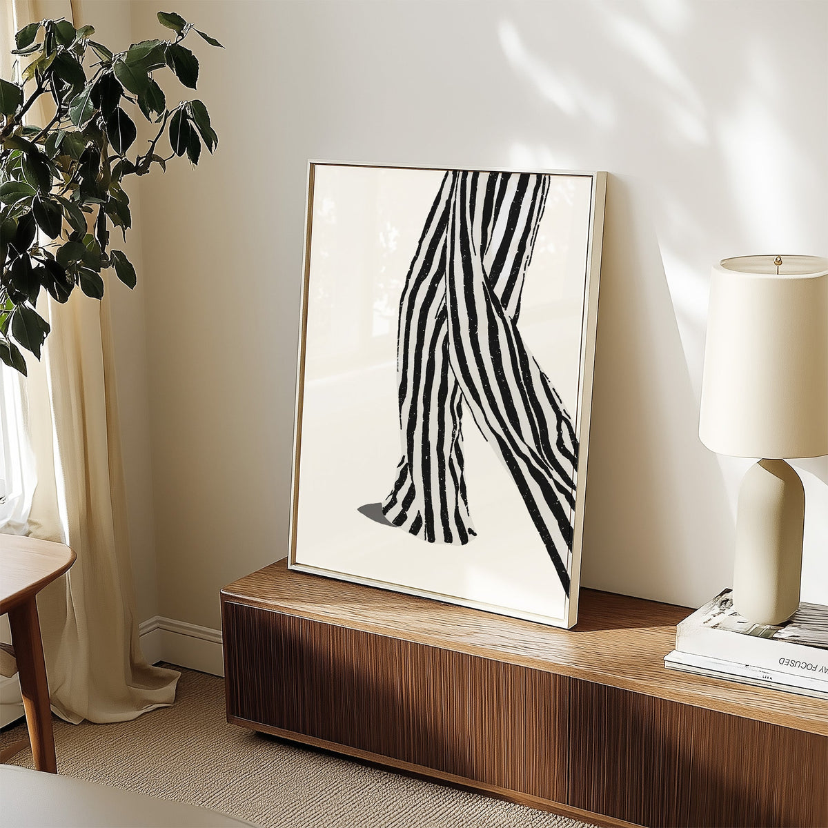 Walking legs in striped trousers graphic print in a white frame on a walnut sideboard beside a ceramic lamp