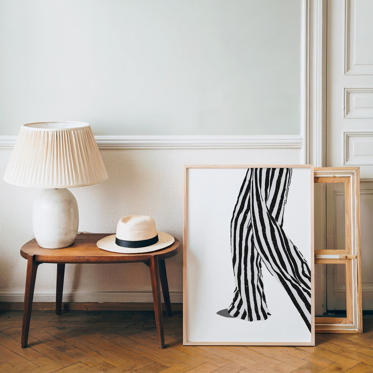 Walking legs in striped trousers graphic print in a pine frame on a wood floor beside a lamp and straw hat