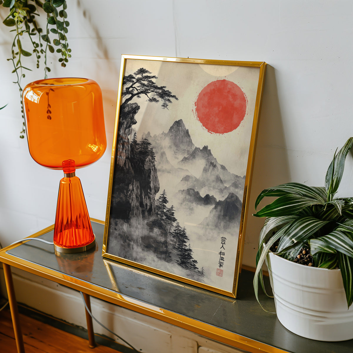 Sumi-e misty mountains and red sun print shown in a gold frame on a console beside an orange glass lamp