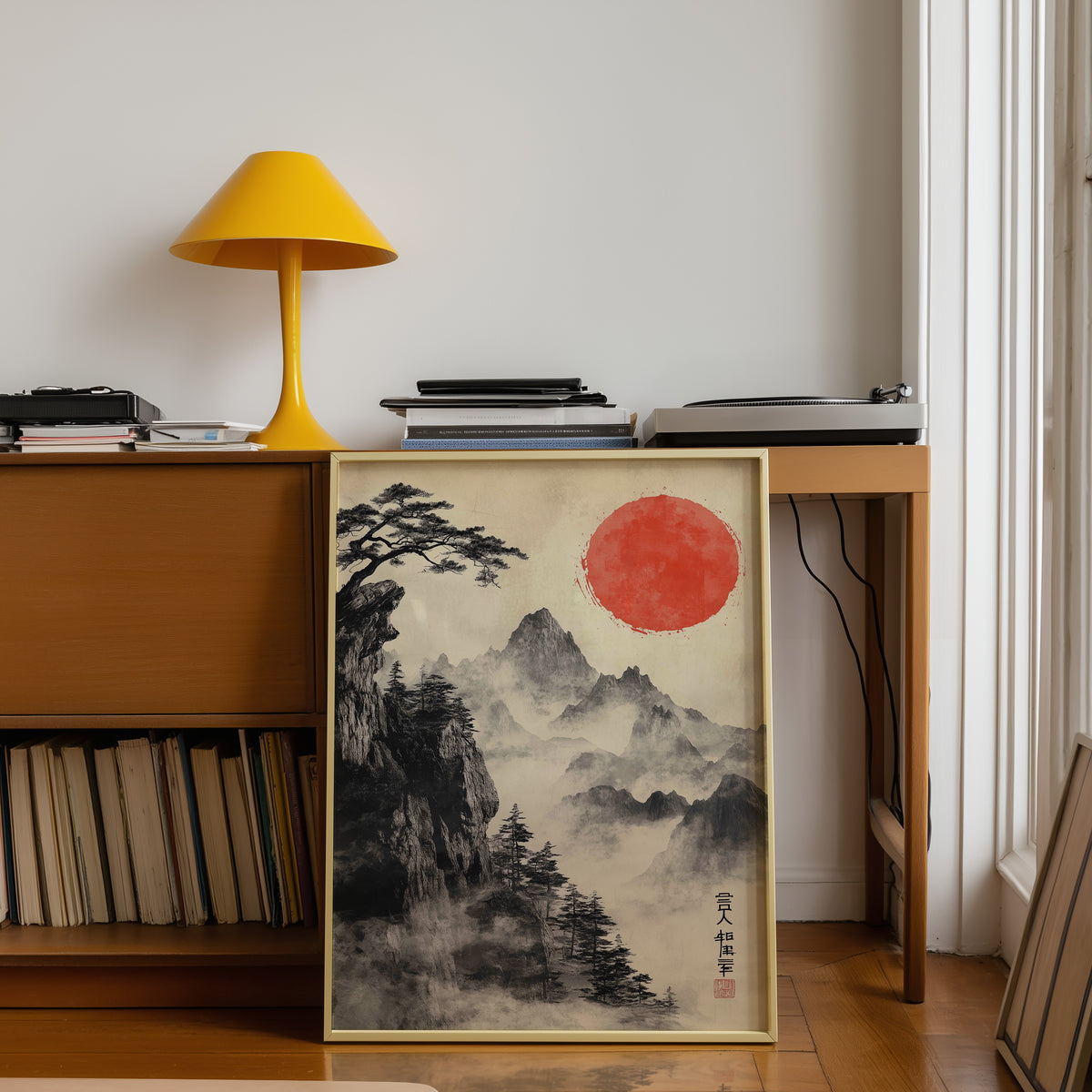 Sumi-e misty mountains and red sun print shown in a gold frame below a teak desk with a yellow lamp