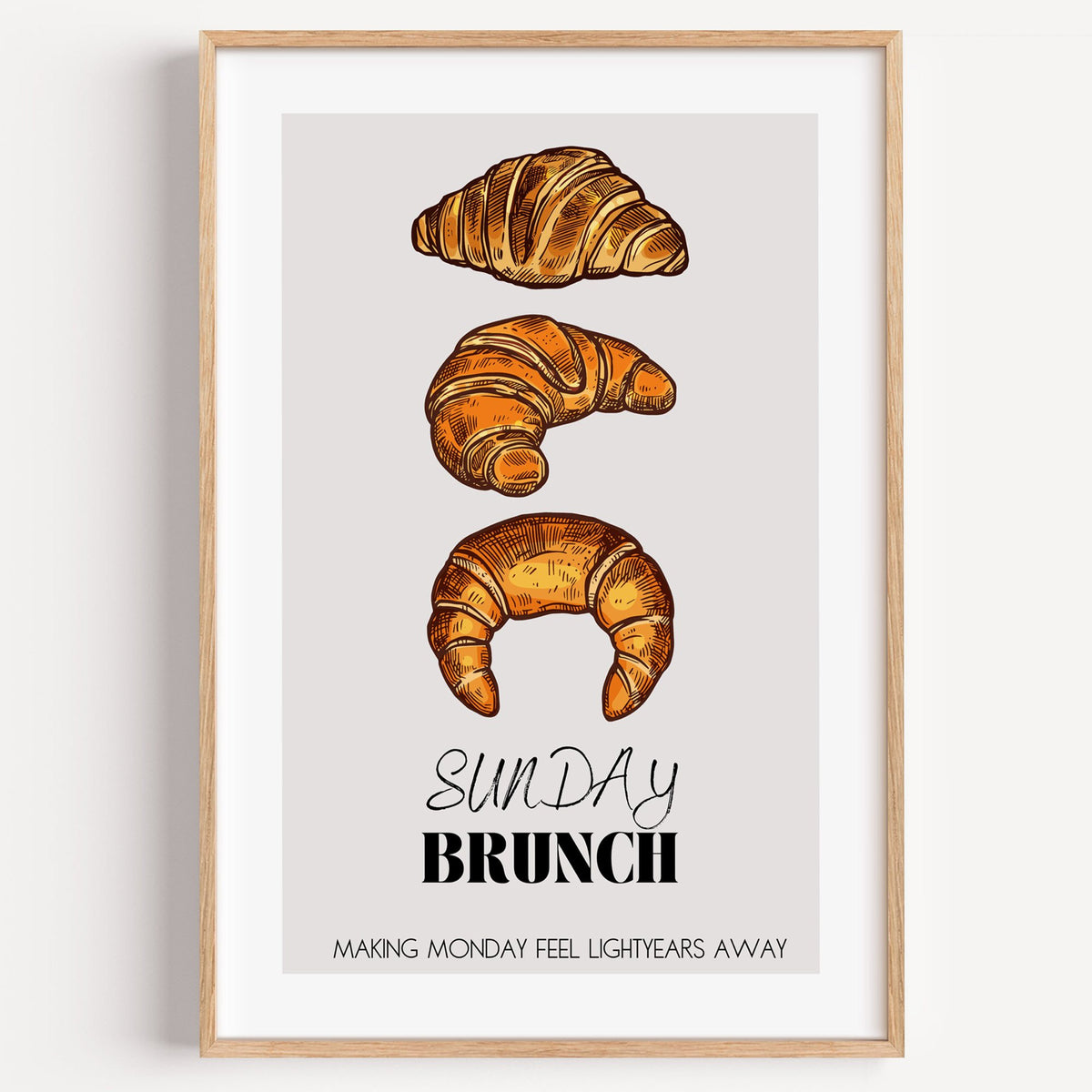 SUNDAY BRUNCH three croissants print in a light oak frame on a plain white wall