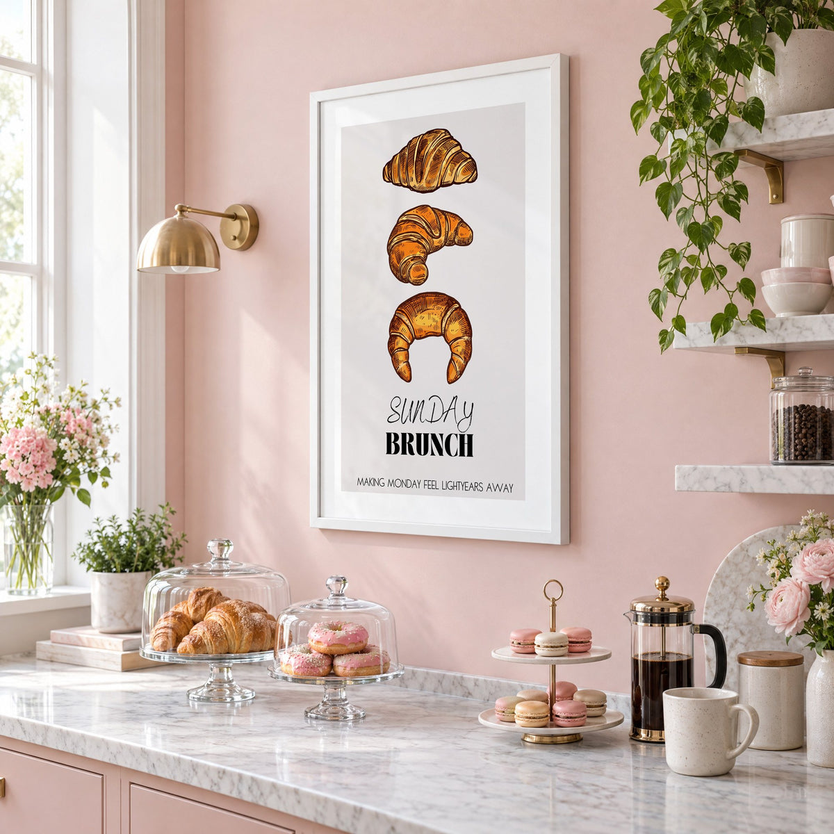 SUNDAY BRUNCH three croissants print in a white frame on a blush pink wall above a patisserie counter with croissants and macarons, seen at an angle