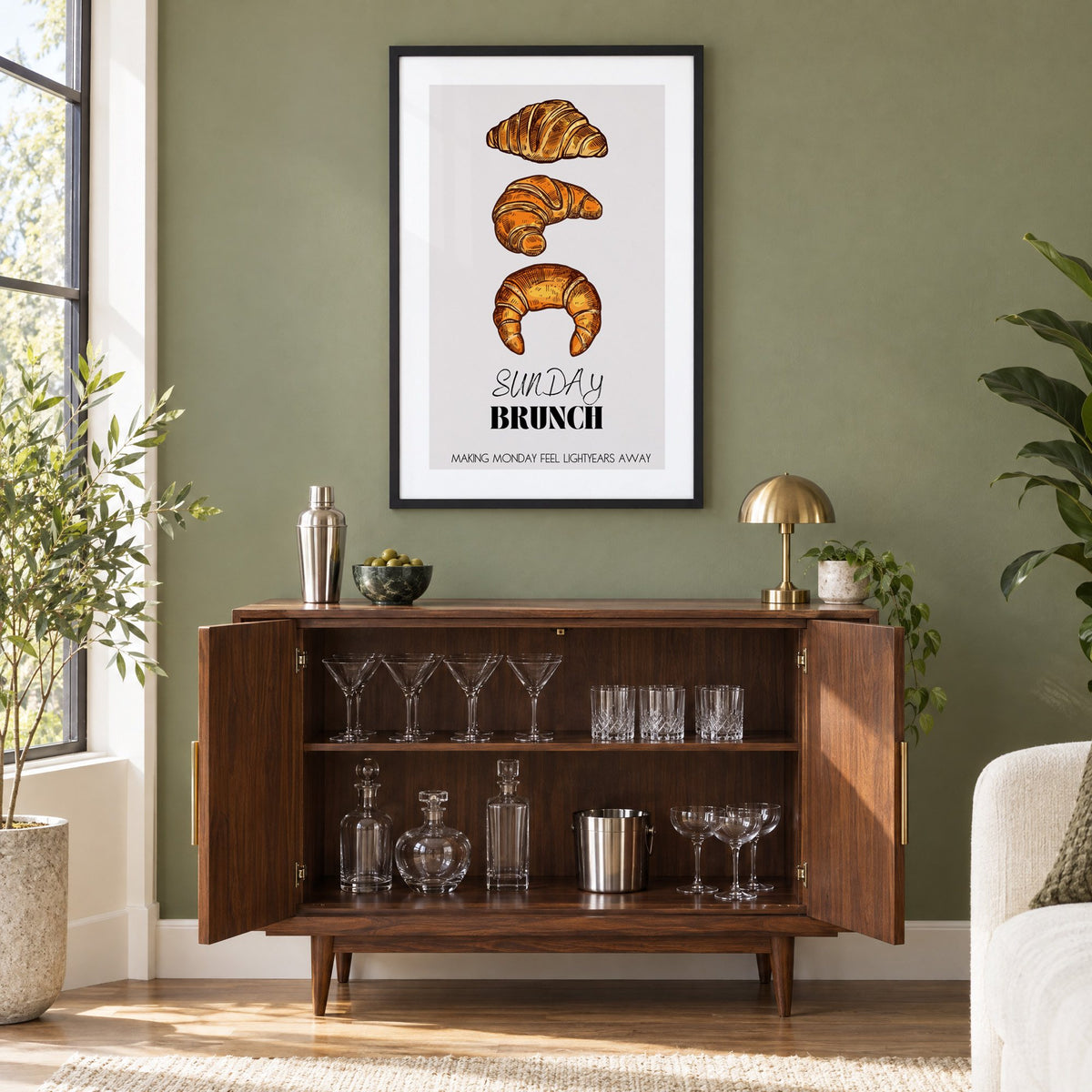 SUNDAY BRUNCH three croissants print in a black frame on an olive green wall above a walnut bar cabinet with glassware