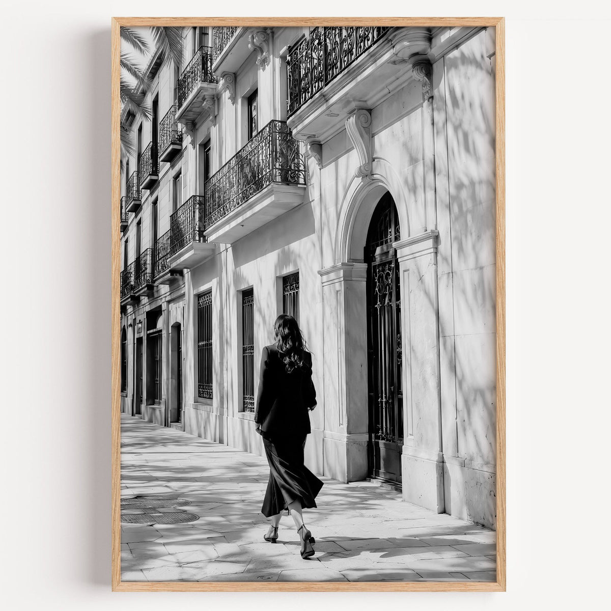 Black and white woman walking under iron balconies print in a light oak frame on a plain white wall