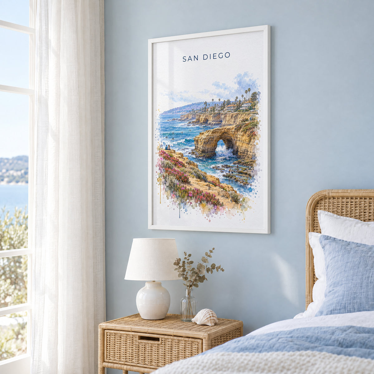 Sunset Cliffs San Diego watercolor print in a white frame on a pale blue wall beside a rattan bed in a coastal bedroom