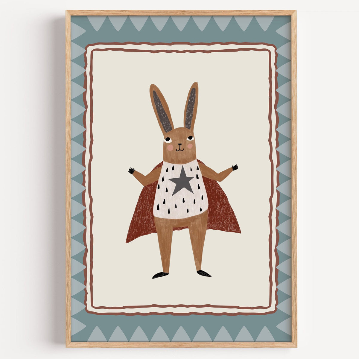 Superhero bunny in red cape nursery print in a light oak frame on a plain white wall