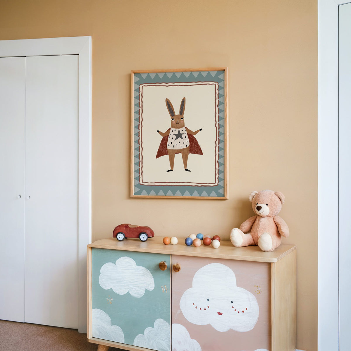 Superhero bunny in red cape nursery print in an oak frame on a peach wall above a cloud-painted cabinet