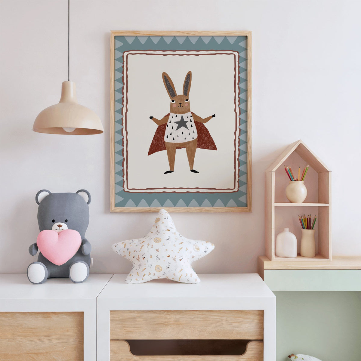 Superhero bunny in red cape nursery print in an oak frame above a white dresser with a star pillow and house shelf