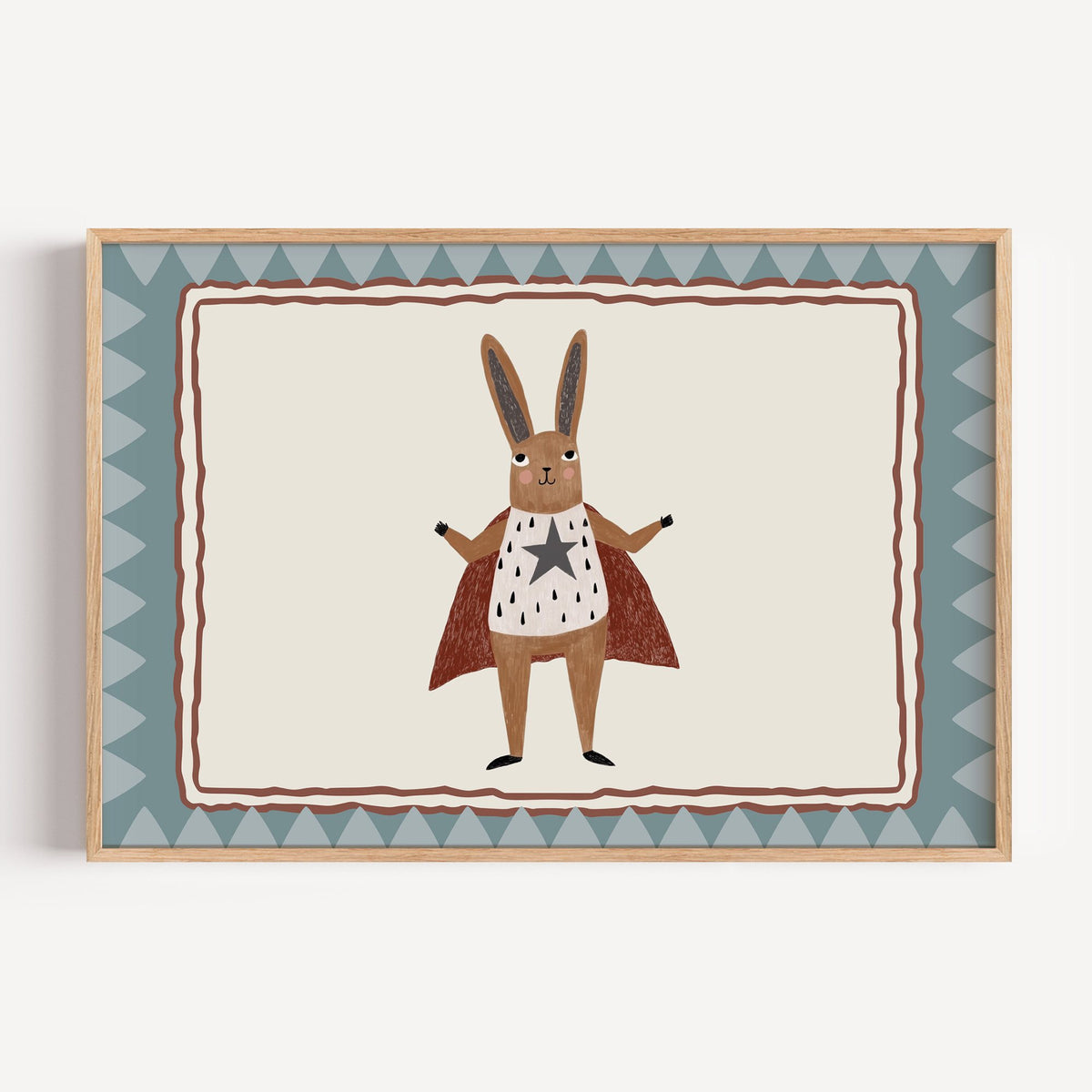 Landscape superhero bunny in red cape nursery print in a light oak frame on a plain white wall