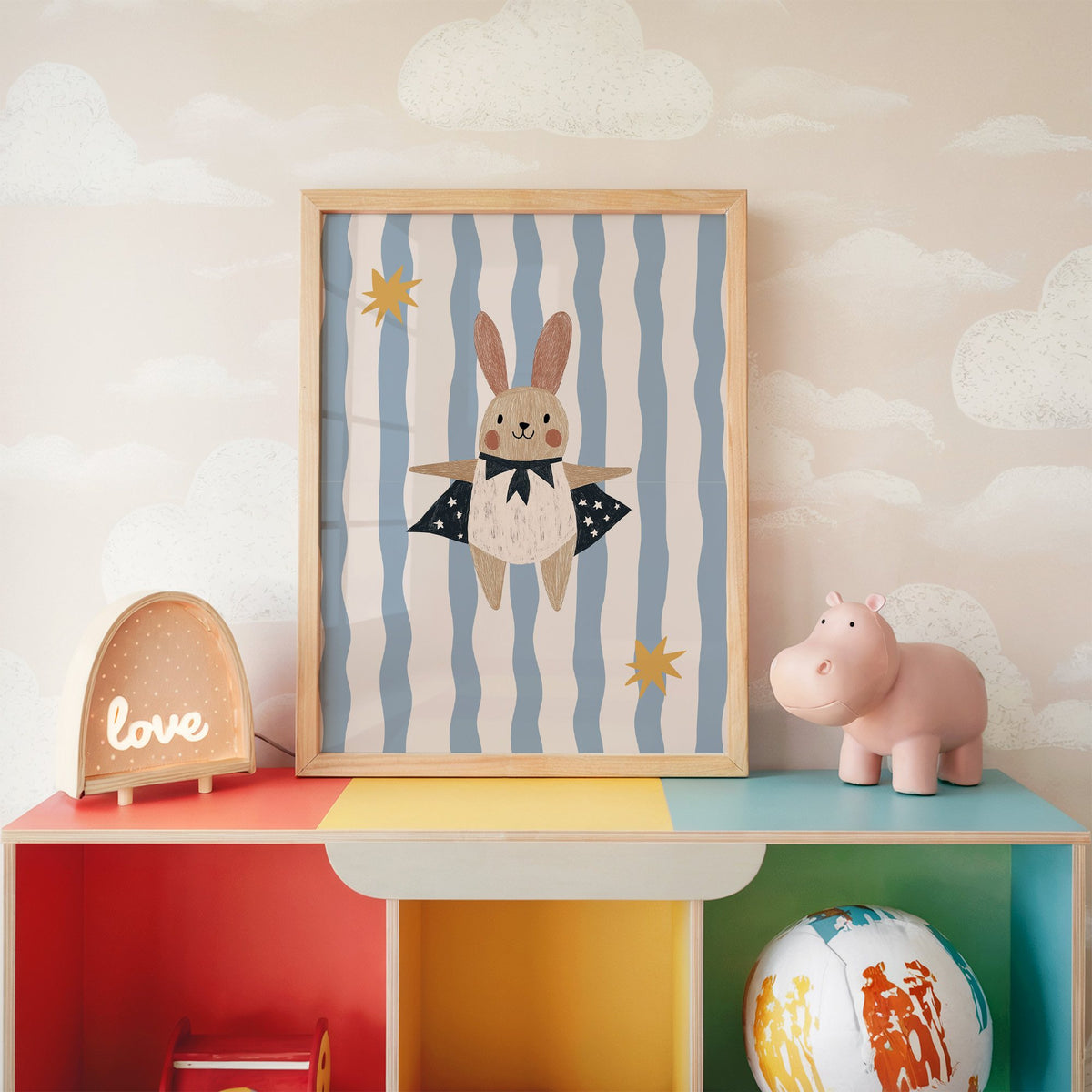 Superhero bunny star cape nursery print in an oak frame on a rainbow toy shelf against cloud wallpaper