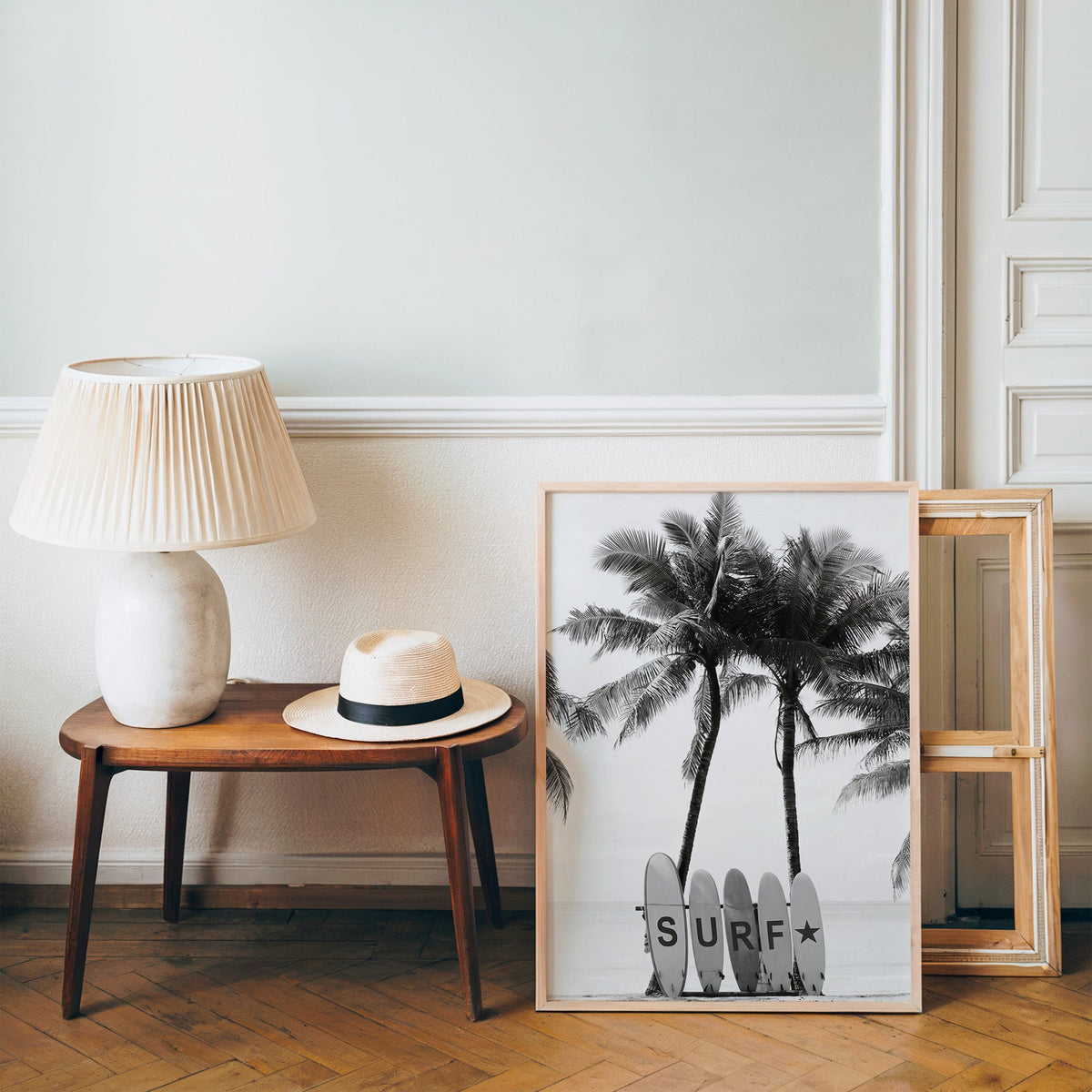 Five surfboards spelling SURF under coconut palms print in a pine frame on a wood floor beside a lamp and straw hat