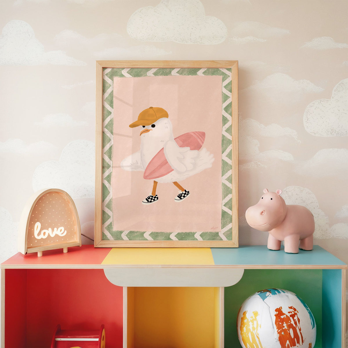 Surfer seagull with pink surfboard nursery print in an oak frame on a rainbow toy shelf against cloud wallpaper