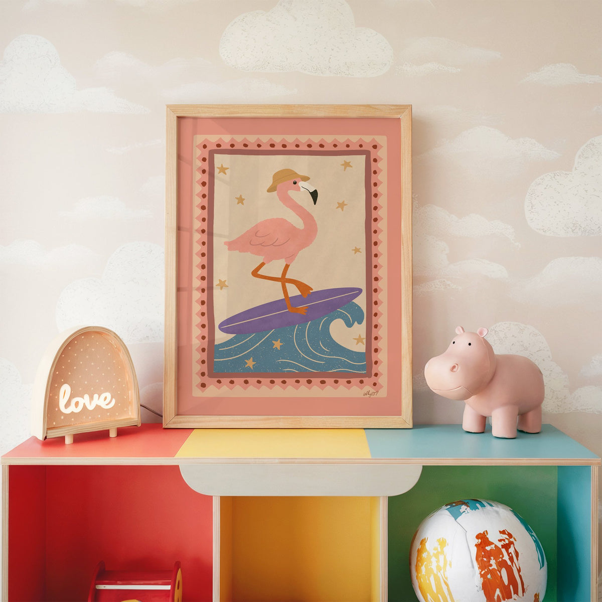 Flamingo on purple surfboard nursery print in an oak frame on a rainbow toy shelf against cloud wallpaper