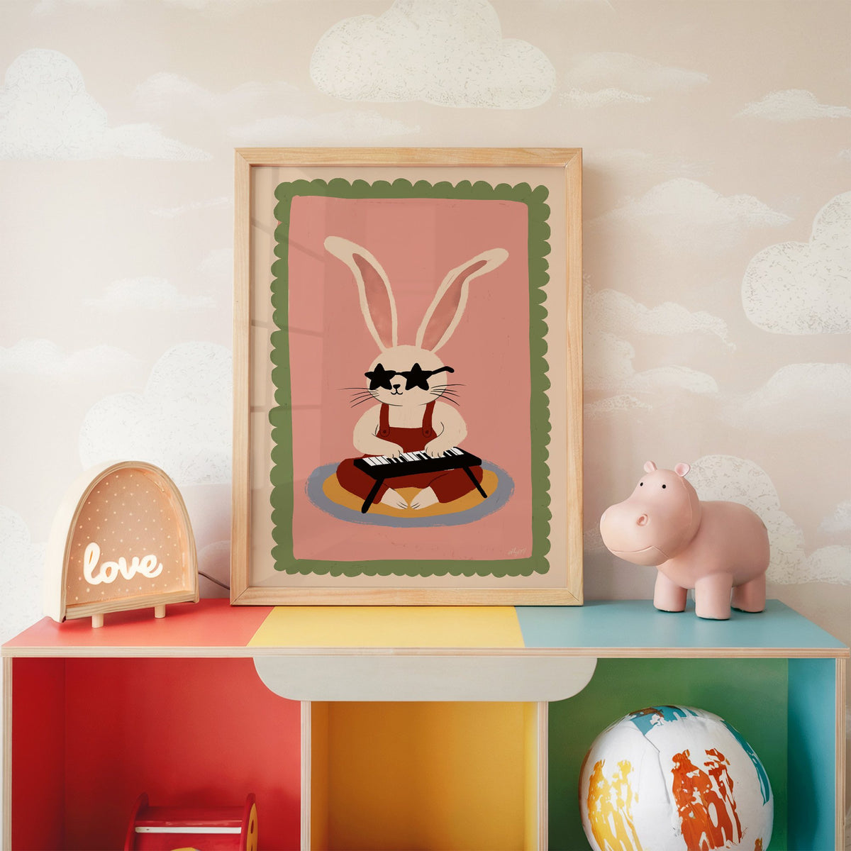 Bunny playing keyboard in star glasses nursery print in an oak frame on a rainbow toy shelf against cloud wallpaper