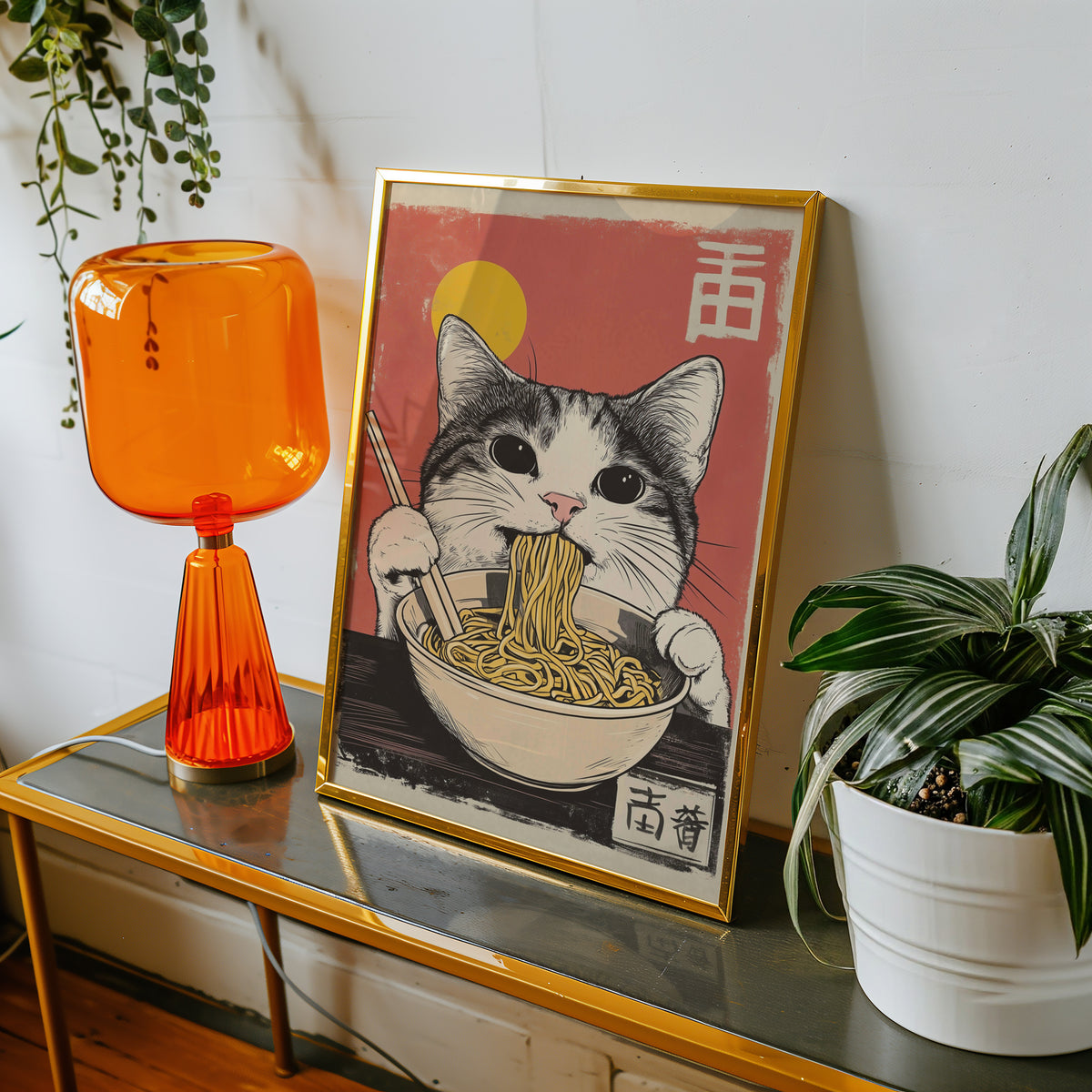 Gray tabby cat eating ramen print shown in a gold frame on a console beside an orange glass lamp