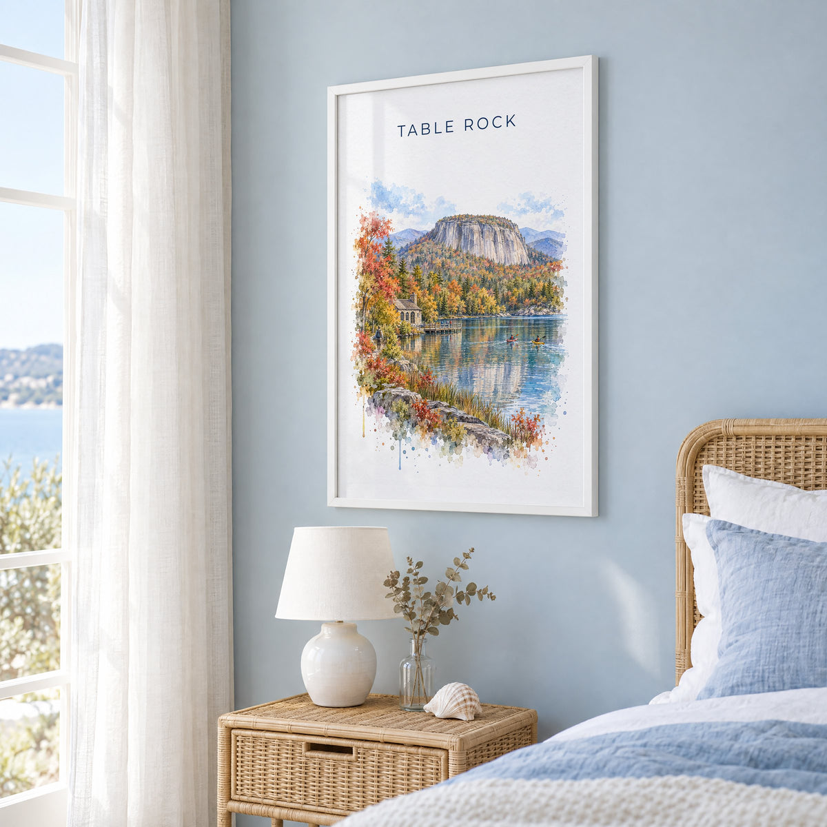 Table Rock State Park watercolor print in a white frame on a pale blue wall beside a rattan bed in a coastal bedroom