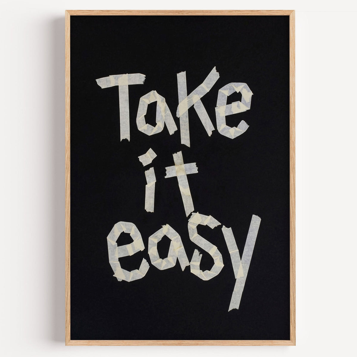 Take It Easy lowercase paper tape letters on black print in a light oak frame on a plain white wall