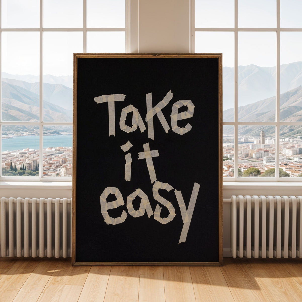 Take It Easy lowercase paper tape letters on black print shown in an antique gold frame by tall windows with a sea view