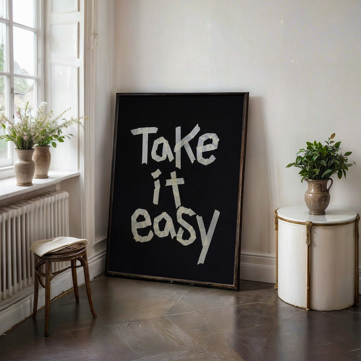 Take It Easy lowercase paper tape letters on black print in a black frame on a dark stone floor beside a white side table