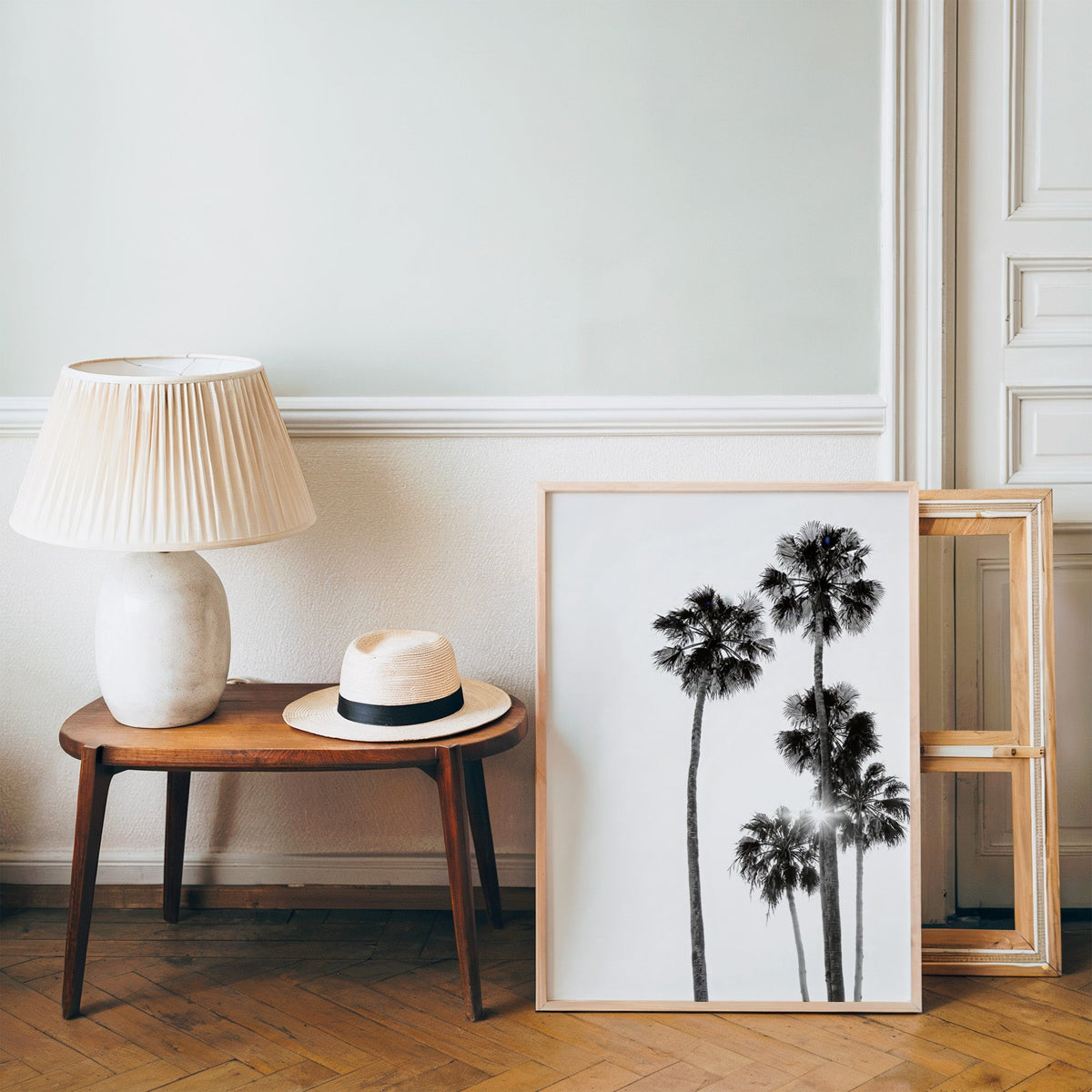 Five tall fan palms with sun flare print in a pine frame on a wood floor beside a lamp and straw hat