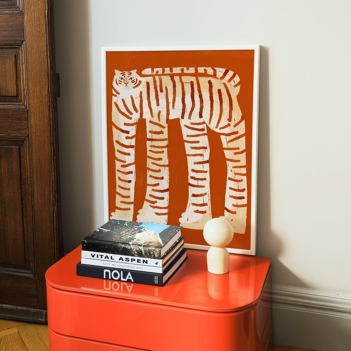 Folk art tiger print in a white frame on a glossy orange cabinet with books